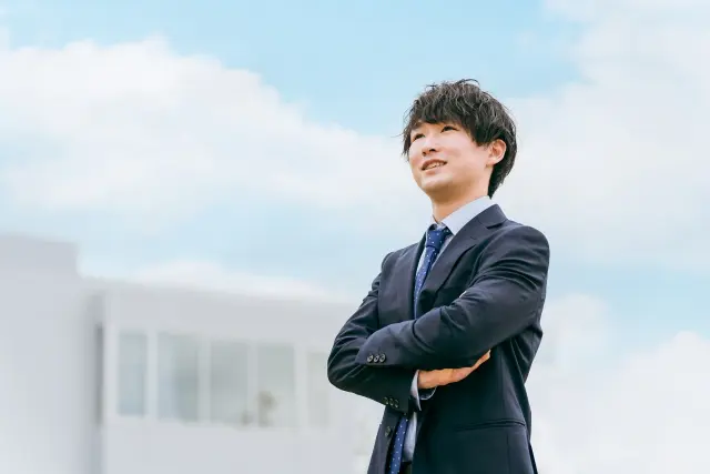 青空をバックに腕を組むスーツ男性
